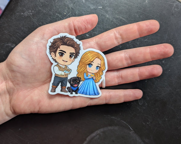 Hand holding a sticker of two animated characters on a dark surface.